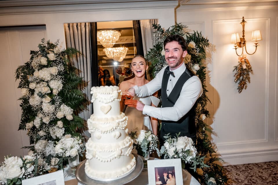 Bride and groom smiling while cutting their wedding cake at an elegant reception, surrounded by floral decor.