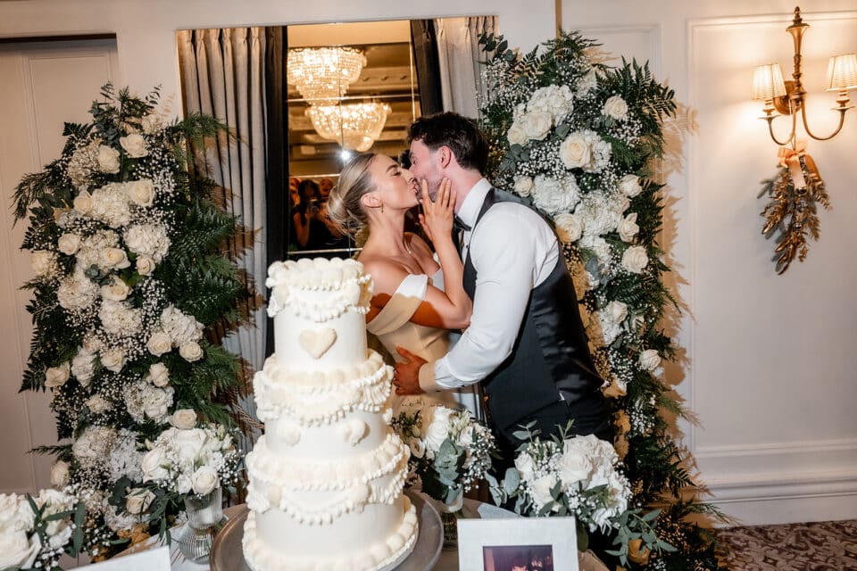 Bride and groom kissing near a wedding cake at Bellingham Castle, surrounded by white floral arrangements.