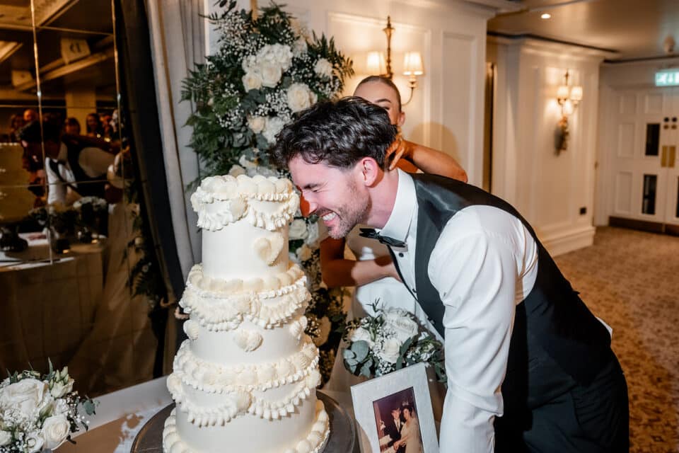 Groom playfully leans towards a tiered wedding cake with bride behind him at a reception in Bellingham Castle.
