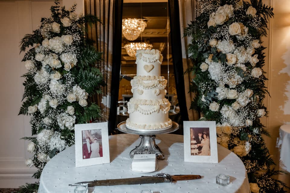 Elegant tiered wedding cake with floral backdrop and framed photos at Bellingham Castle.
