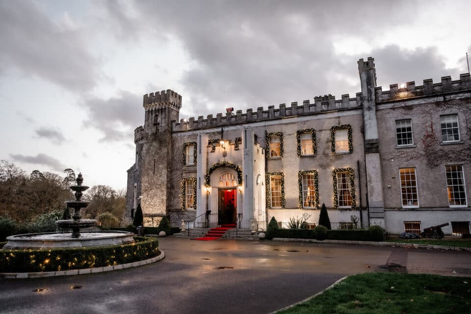 Bellingham Castle entrance decorated with lights and a red carpet, with a fountain in the foreground and cloudy sky.