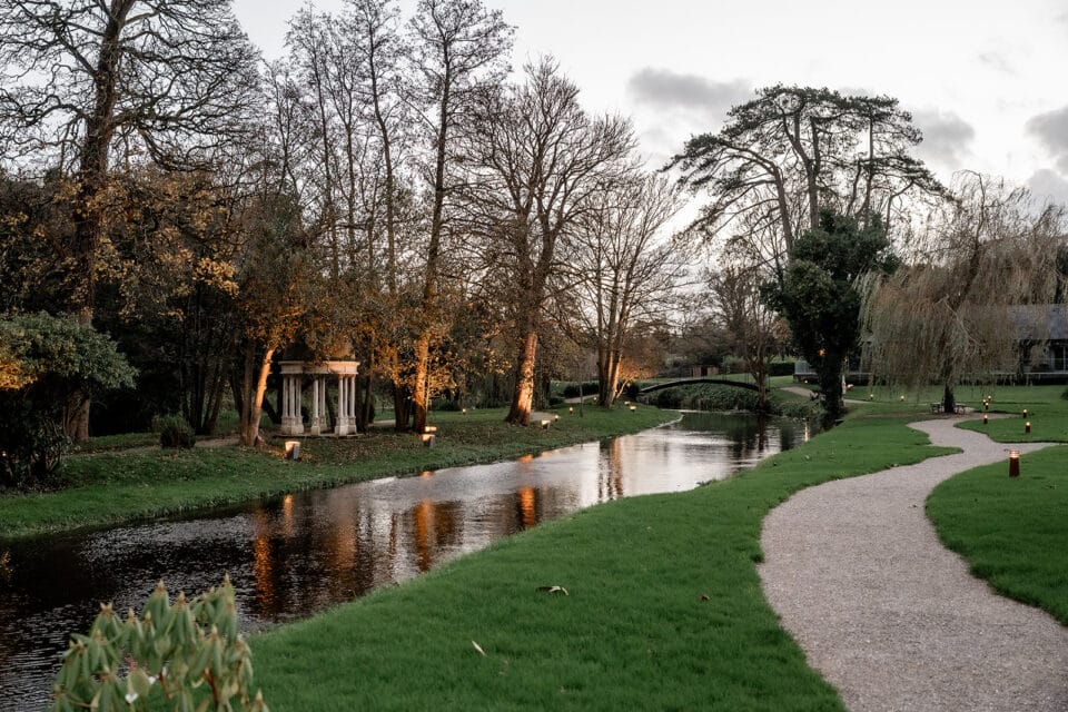 Peaceful garden scene at Bellingham Castle with a winding path, stream, and autumn trees.