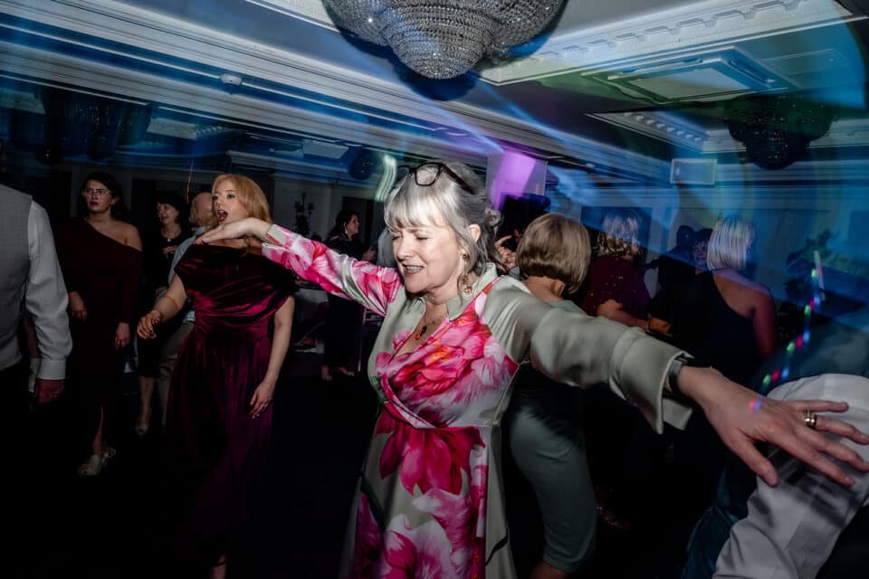 Woman dancing energetically at a lively indoor event with colorful lighting and other guests in the background.