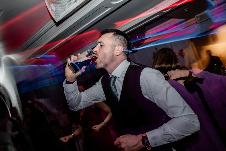 Man in formal attire drinking from a pint glass at a lively indoor event.