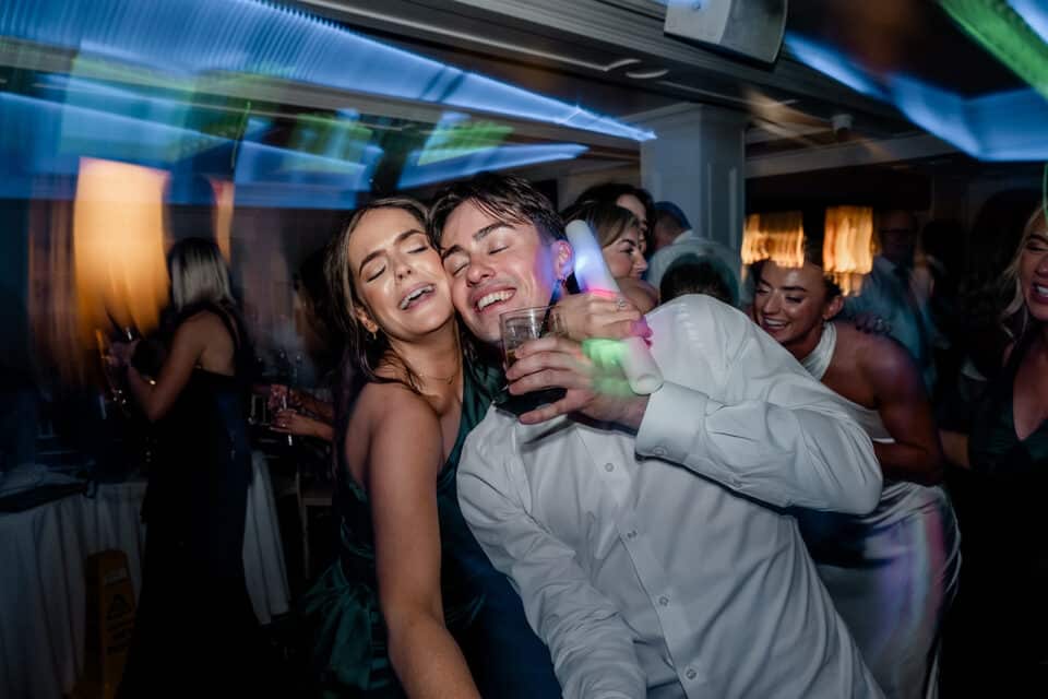 A joyful couple dancing with friends at a party, holding drinks and glow sticks, at an indoor celebration.