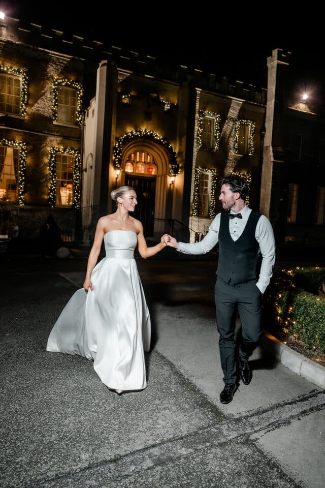 Bride and groom holding hands in front of Bellingham Castle at night.