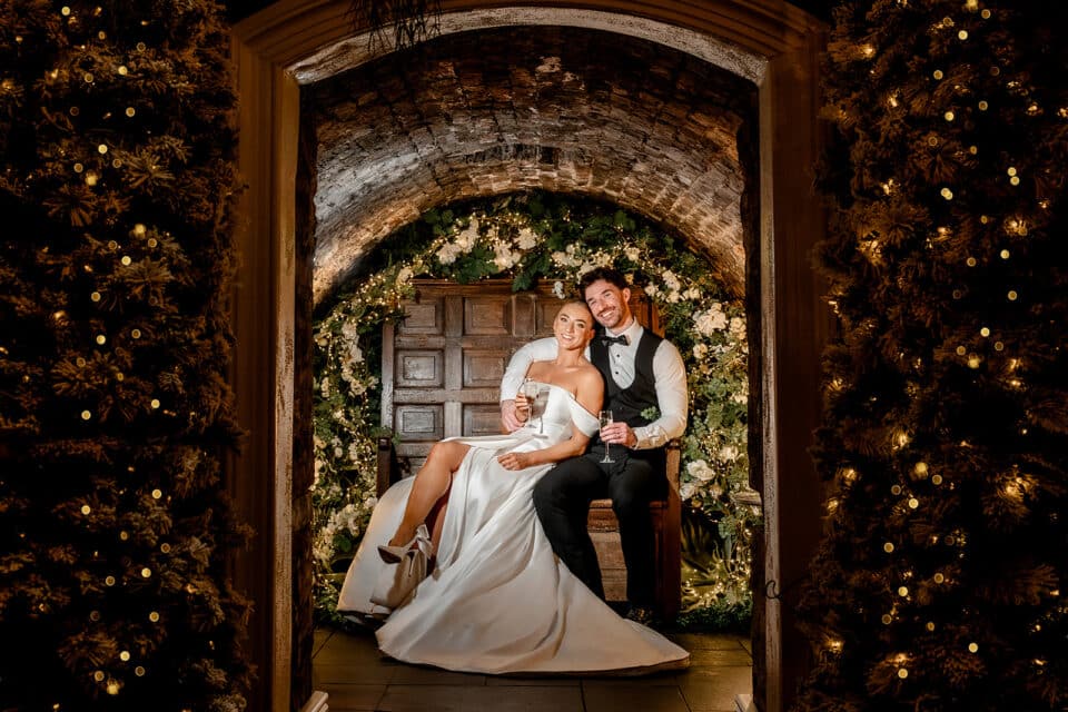 Bride and groom sitting in an archway decorated with flowers and lights at Bellingham Castle.
