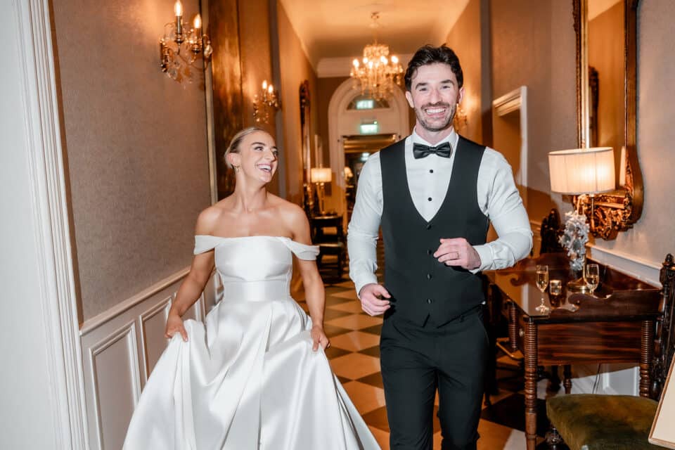 Bride and groom smiling as they walk through an elegantly decorated hallway at Bellingham Castle.