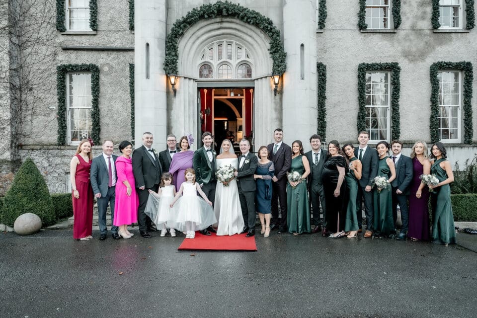 Wedding party in formal attire outside Bellingham Castle, with bride and groom in the center, surrounded by family and friends.