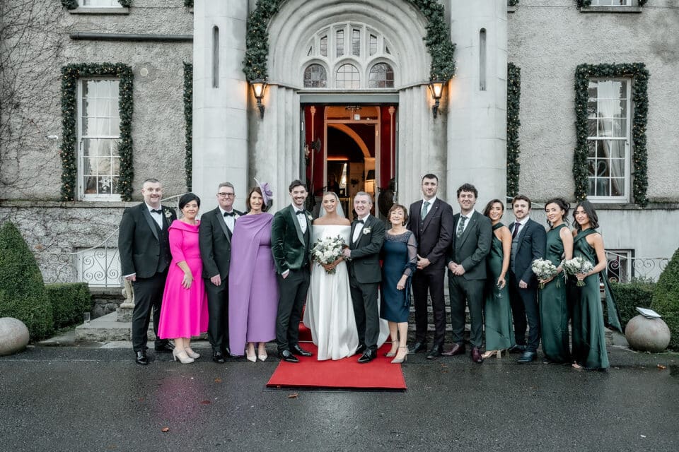 Wedding party poses outside Bellingham Castle, featuring bride, groom, and guests in formal attire on a red carpet.