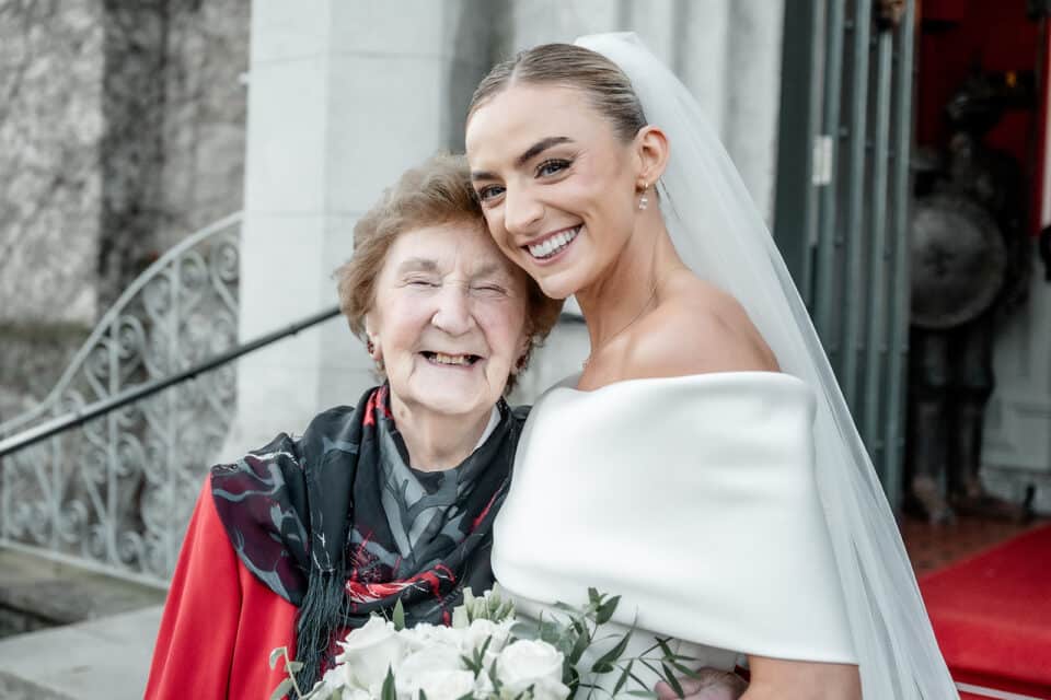 Bride smiling with an elderly woman at a wedding, holding a bouquet, outside a building with stone steps.