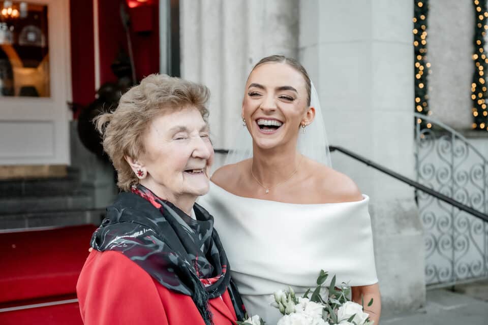 Bride smiling with an elderly woman outside Bellingham Castle, holding a bouquet of white flowers.