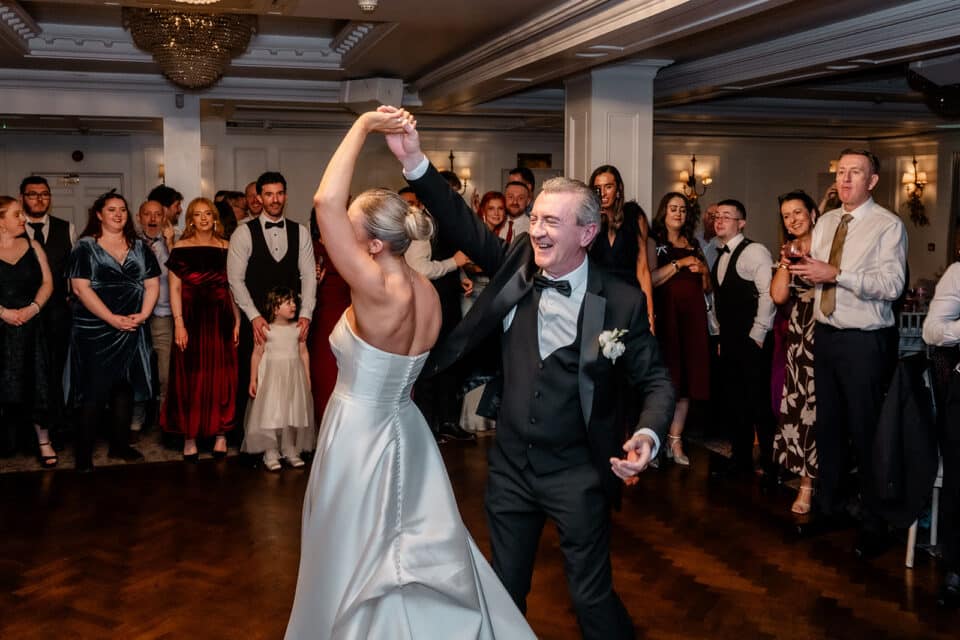 Bride and groom dancing at their wedding reception with guests watching in a decorated hall.