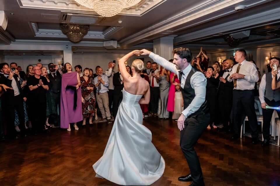 Bride and groom dancing at wedding reception, surrounded by applauding guests in elegant attire at Bellingham Castle.