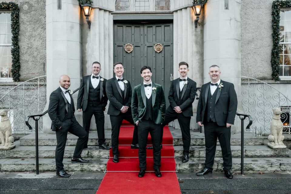 Groomsmen in formal suits pose on red carpet at Bellingham Castle entrance.