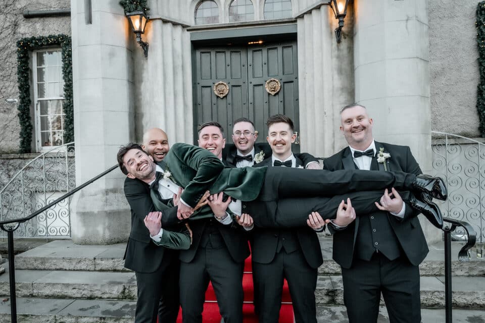 Groomsmen in suits joyfully holding one of their friends outside Bellingham Castle.