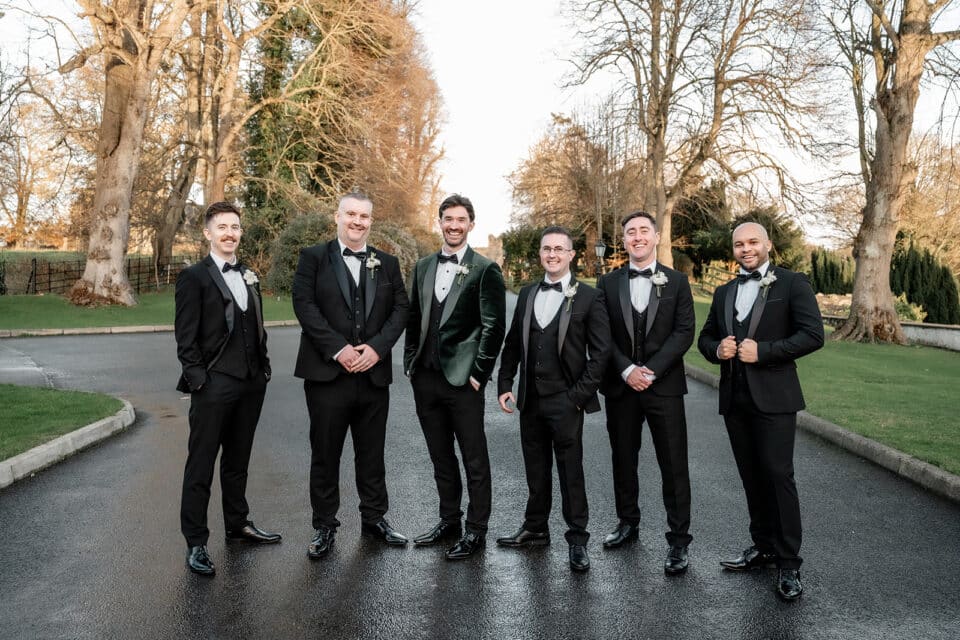Groomsmen in black suits pose outdoors on a paved path at Bellingham Castle.