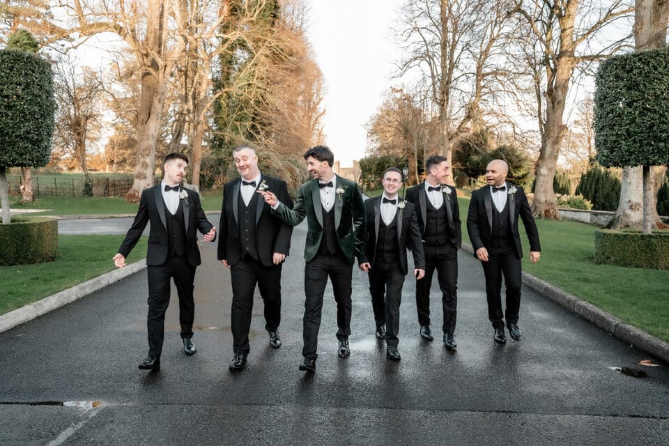 Groomsmen walking and laughing in tuxedos on a tree-lined path at Bellingham Castle.