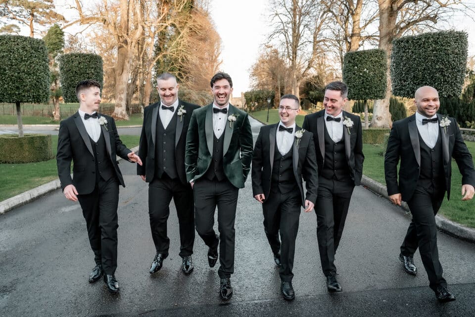 Groomsmen in tuxedos walking at Bellingham Castle grounds, smiling and chatting.