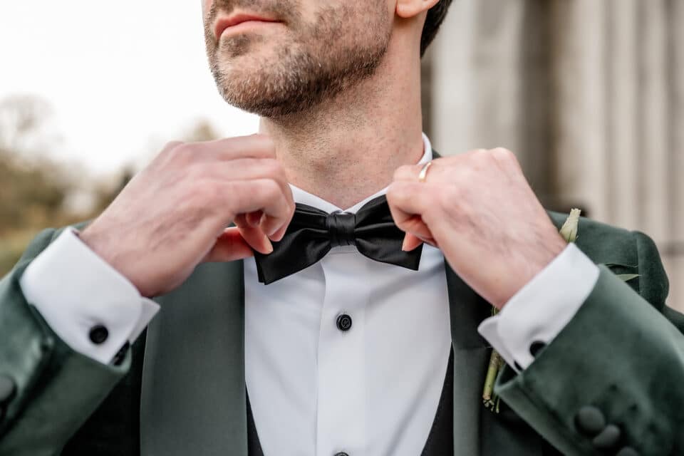 Groom adjusting a black bow tie, wearing a green suit and white shirt, at Bellingham Castle.