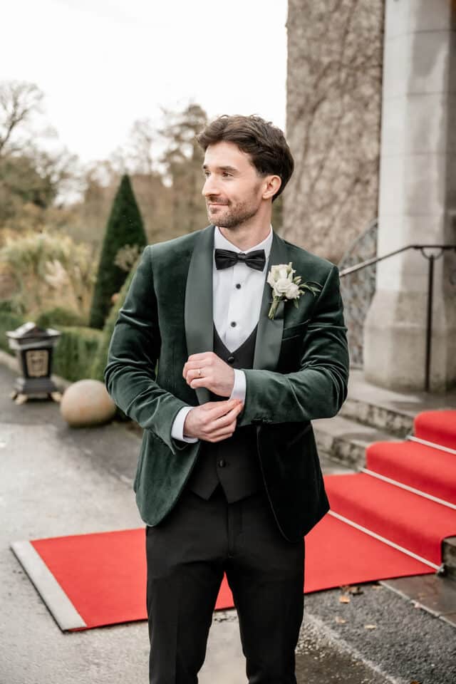 Groom in a green velvet suit adjusts his cufflinks on a red carpet outside a castle.