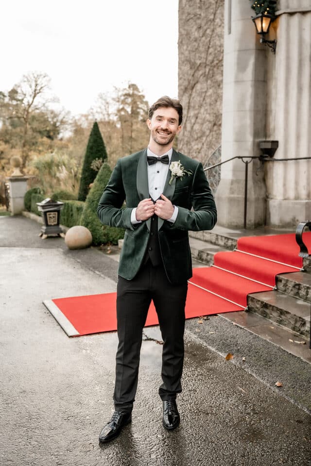Man in a green tuxedo stands smiling on a red carpet outside a castle entrance.