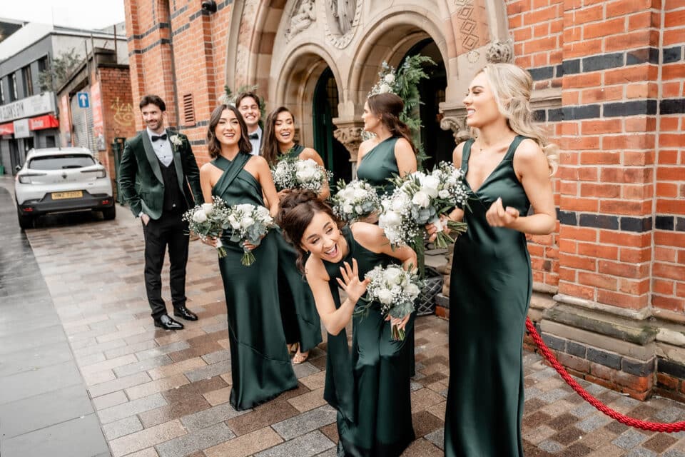 Bridesmaids in green dresses smile and hold bouquets outside a red brick building on a rainy day.
