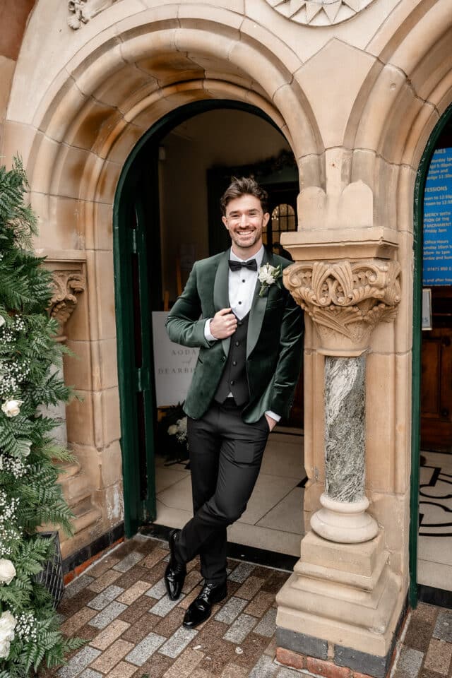 Man in green velvet suit standing at ornate entrance of Bellingham Castle, smiling with hands in pockets.