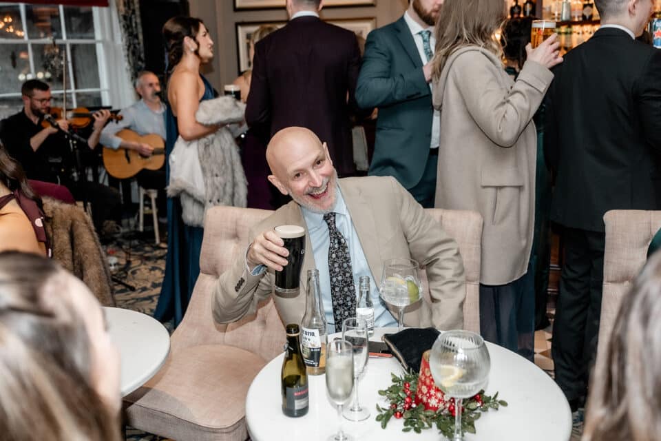 Man in a beige suit holding a pint in a lively bar setting with people mingling and musicians playing in the background.
