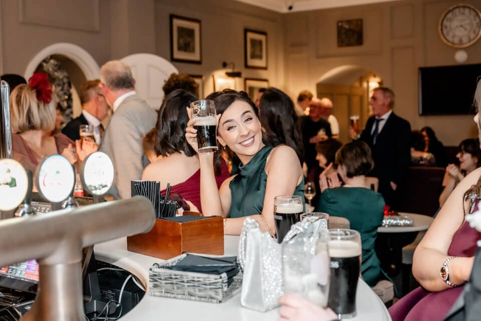 Woman at bar smiling and holding a pint in a lively social gathering at Bellingham Castle.