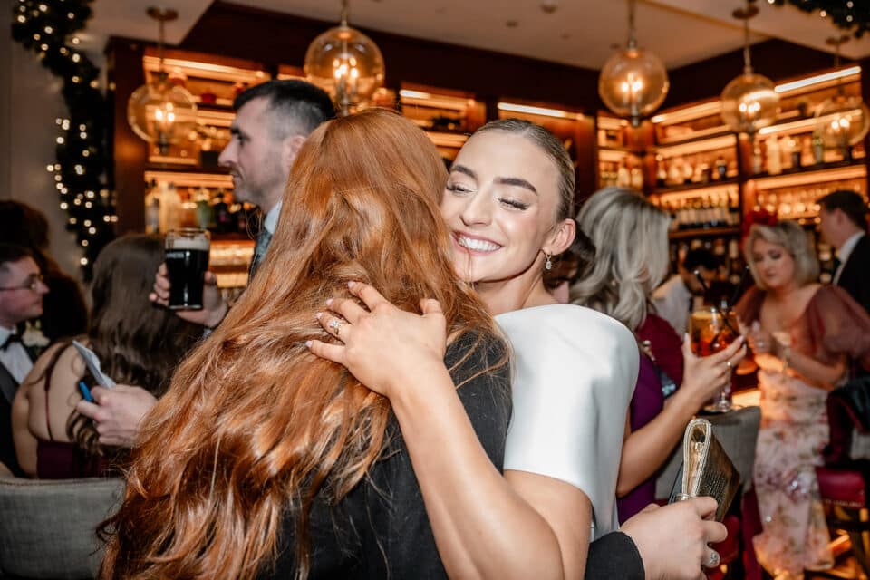 Woman in white dress hugging a person with long red hair at a festive event, surrounded by people in a warmly lit bar.