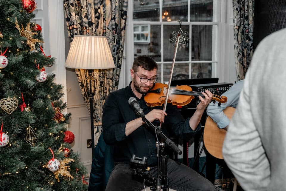 Musician playing violin beside decorated Christmas tree indoors.
