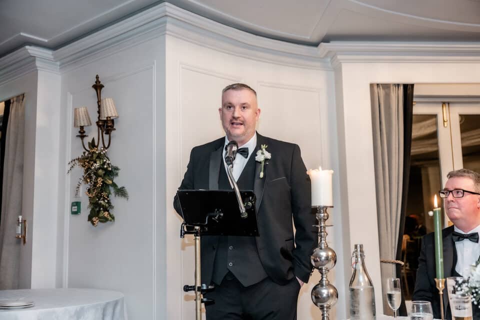 Man in a tuxedo giving a speech at a wedding reception in Bellingham Castle, with a microphone and decorated candles.