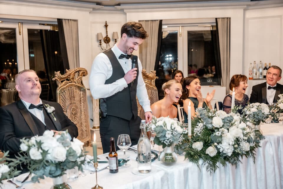 Groom giving speech at wedding reception, guests laughing, floral decorations.