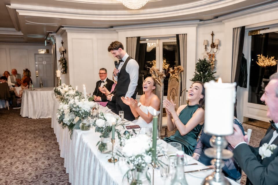 Wedding reception speech at Bellingham Castle, guests seated and clapping, floral decorations on the table.