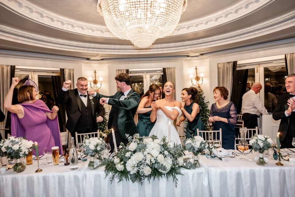 Wedding reception celebration at Bellingham Castle with guests cheering and smiling under a grand chandelier.
