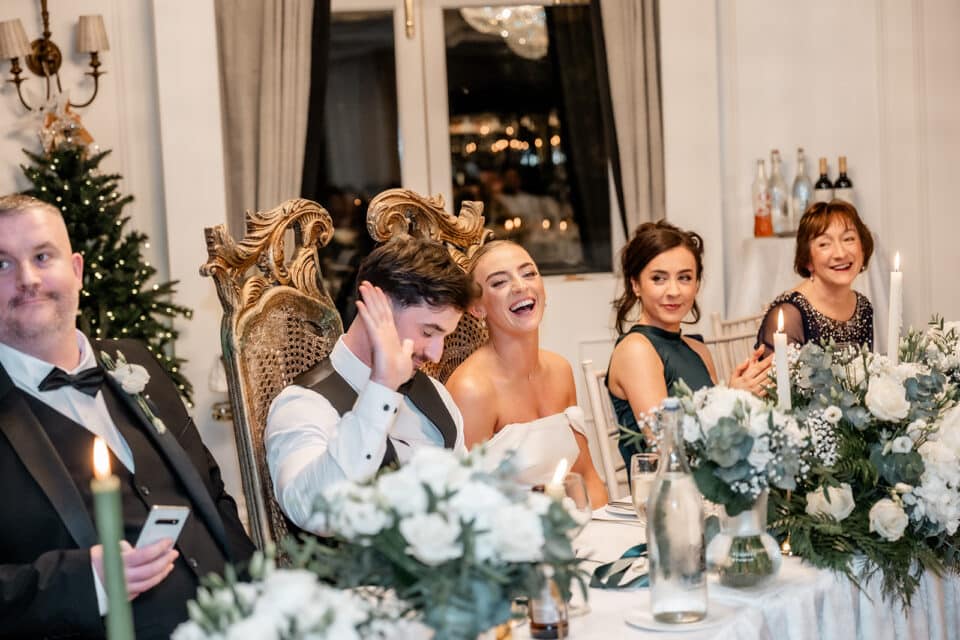 Wedding party at Bellingham Castle, guests laughing and seated at a decorated table with candles and flowers.