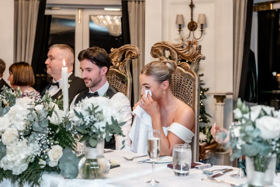 Bride and groom seated at a reception table, bride wiping tears with a napkin, elegant decor and flowers around.