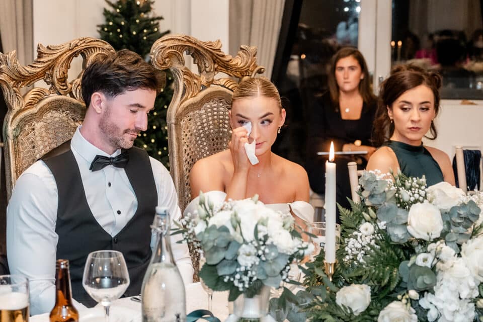 Wedding couple at reception table, bride wiping tears, surrounded by flowers and candles, elegant setting.