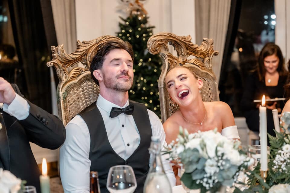 Couple laughing joyfully at a wedding reception with elegant decor and a Christmas tree in the background.