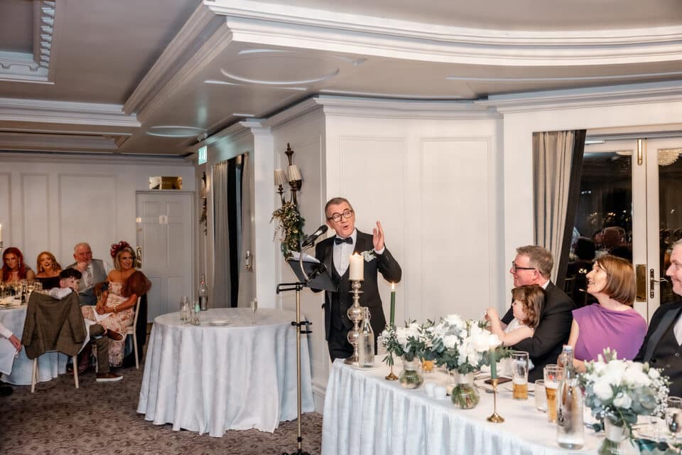 A man giving a speech at a wedding reception in an elegantly decorated room, with guests seated and listening attentively.