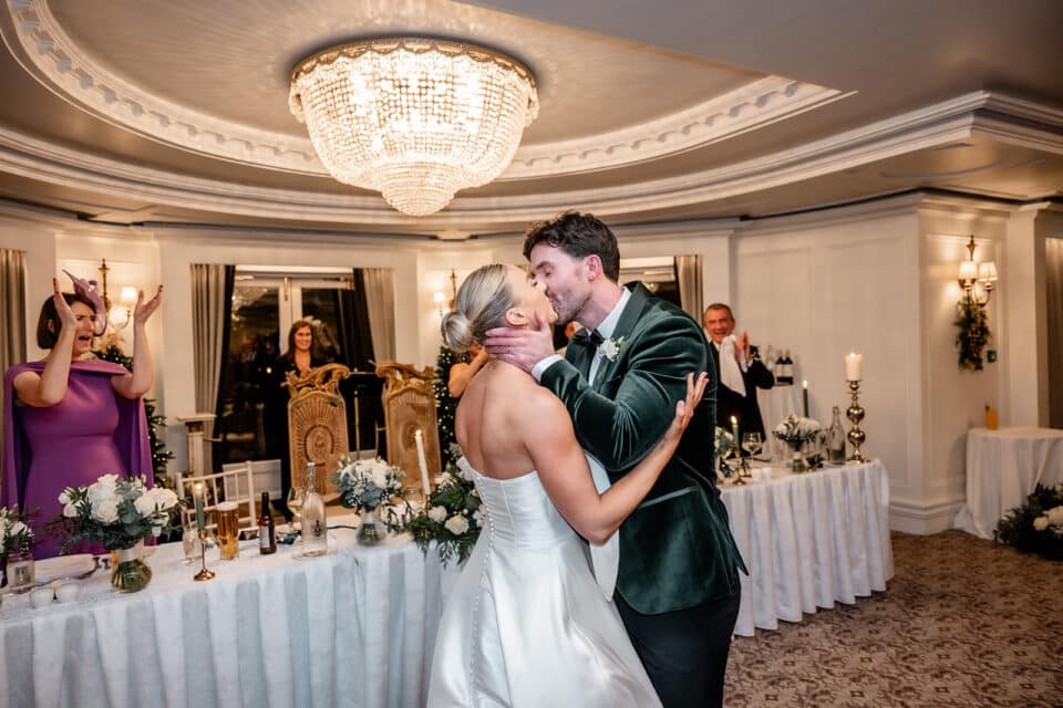 Bride and groom share a kiss at their wedding reception in Bellingham Castle, with guests applauding.