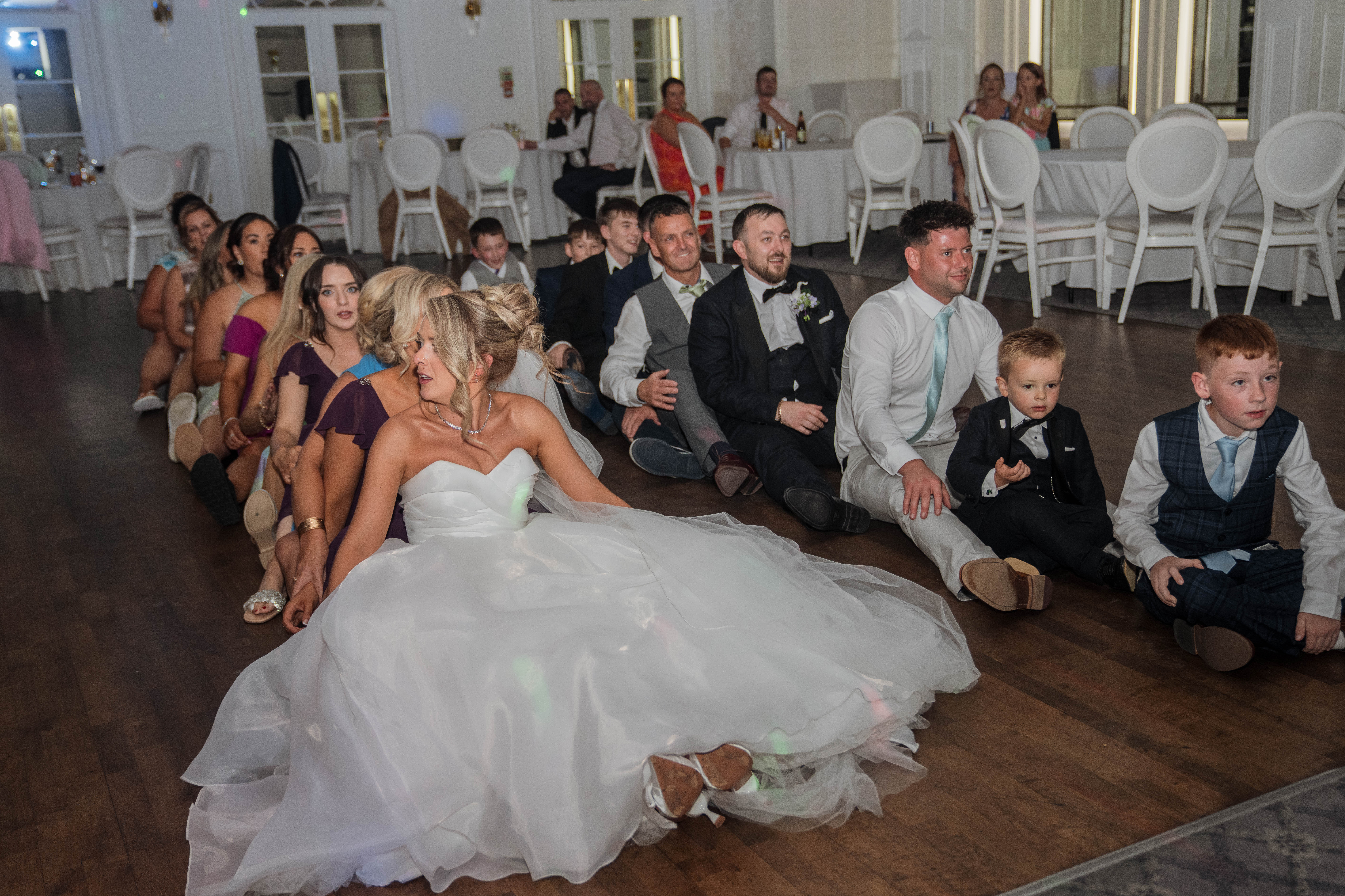 Wedding guests sitting on the floor in a conga line at a reception.