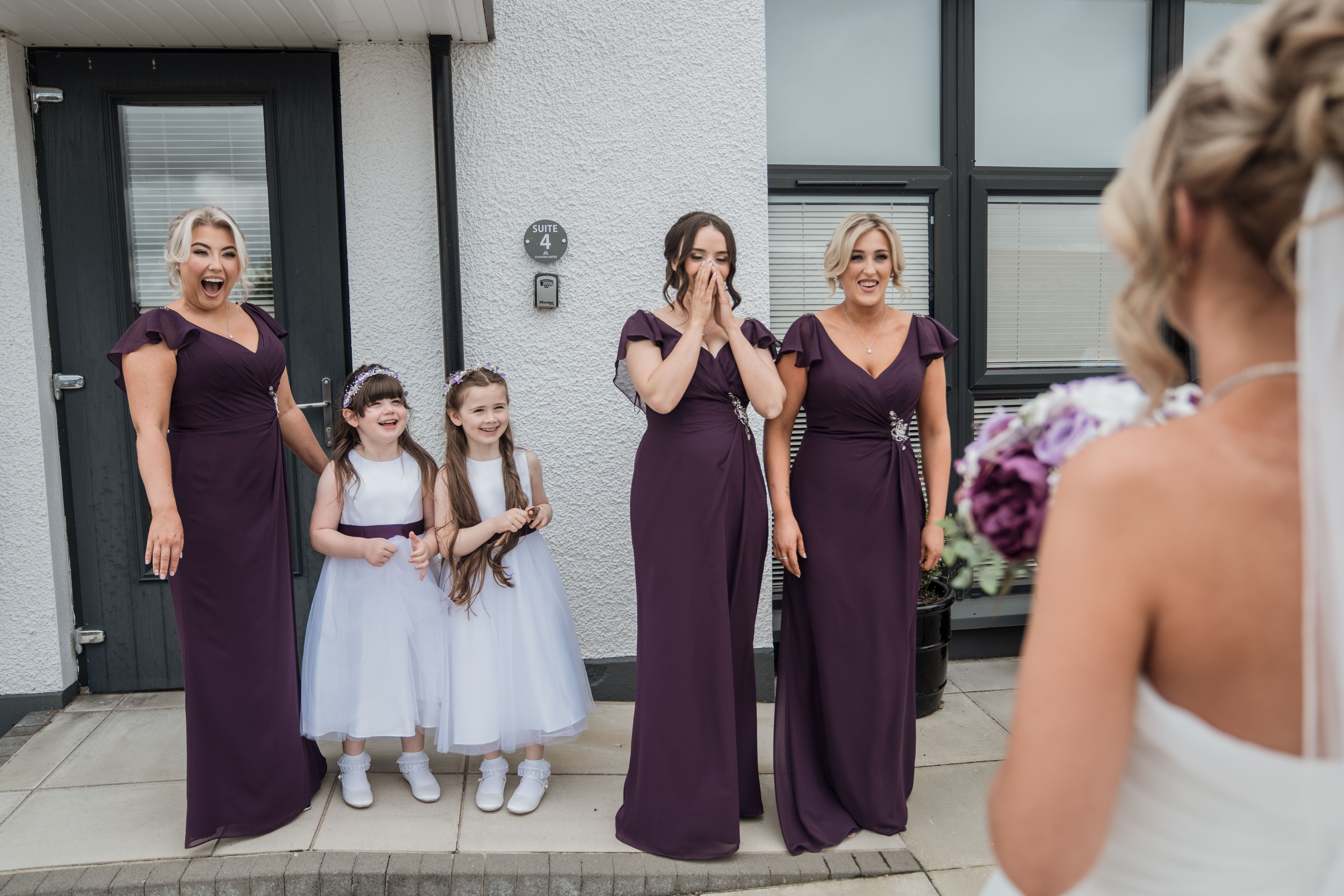 Bridesmaids and flower girls in purple and white dresses react to bride outside a house.