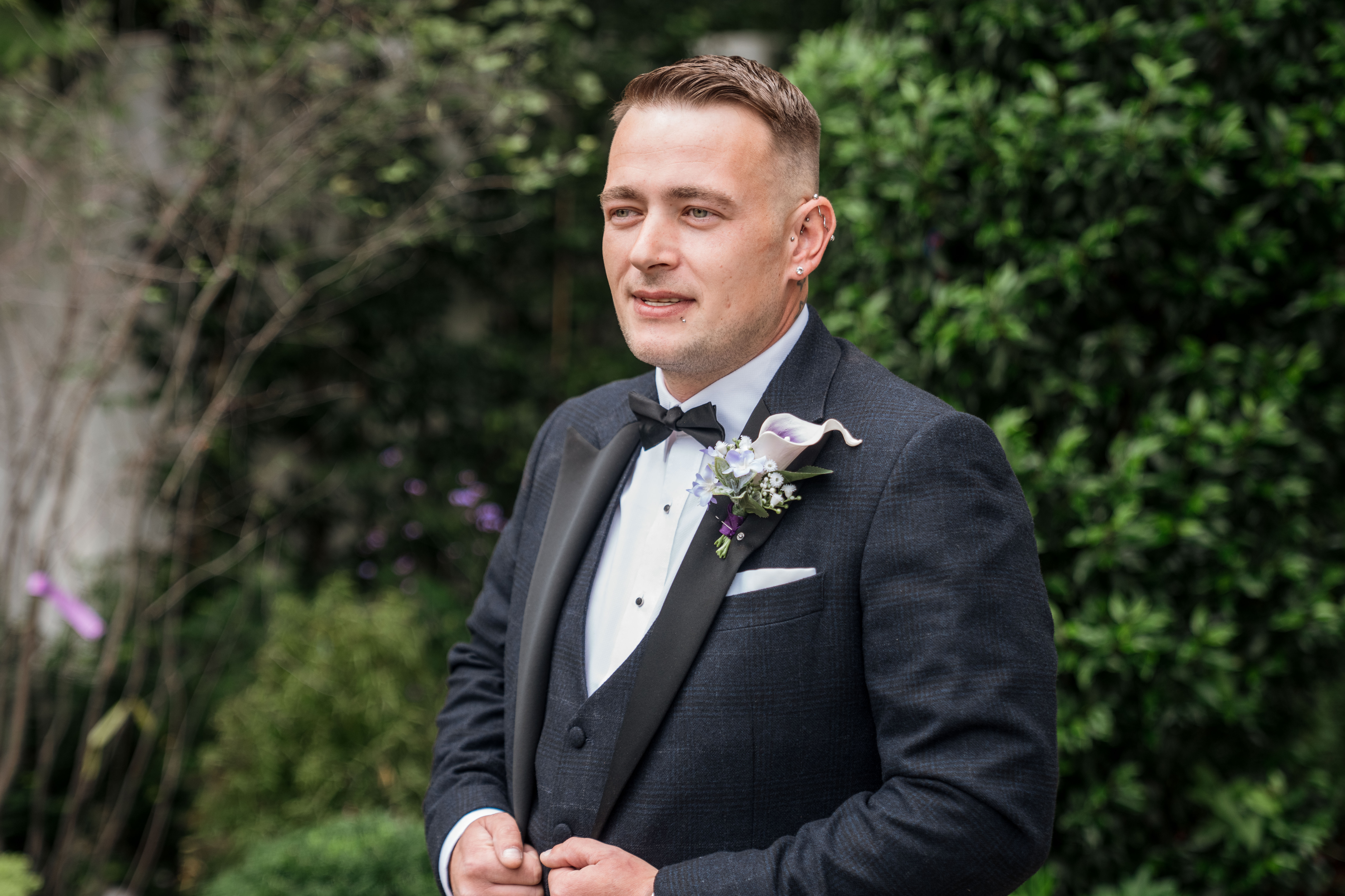 Man in a tuxedo with a boutonniere, standing outdoors with greenery in the background.