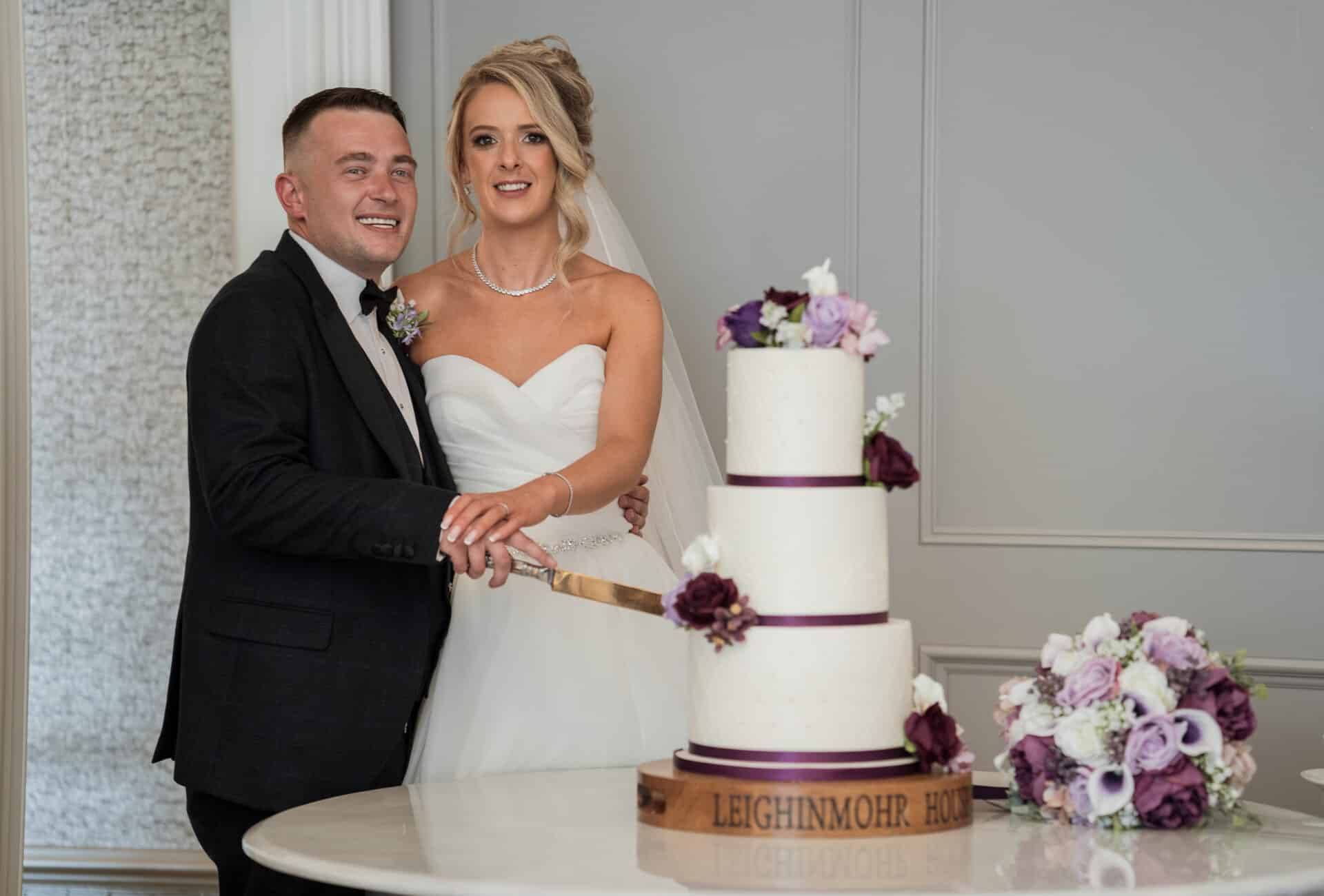 Bride and groom cutting a three-tier wedding cake adorned with purple flowers.