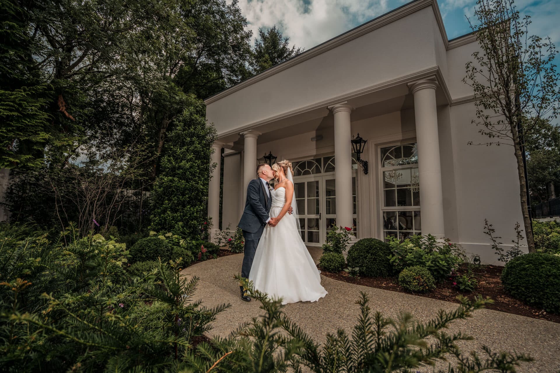 Bride and groom kissing in front of an elegant white building with columns, surrounded by lush greenery.
