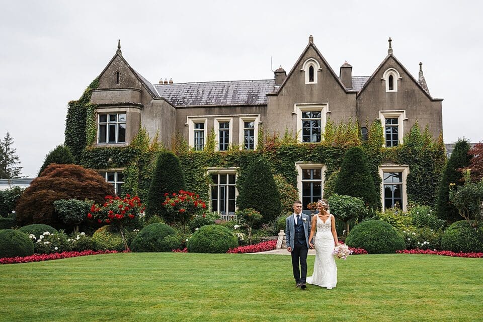 Bride and groom walking on lawn in front of ivy-clad Ballymascanlon Hotel.