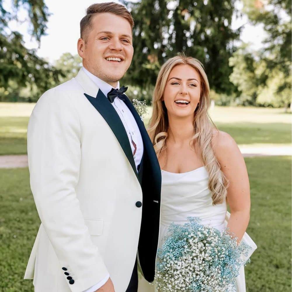 Smiling couple in wedding attire standing outdoors with greenery in the background.