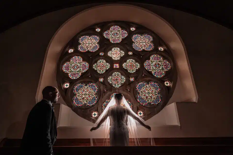 Bride standing beneath stained glass windows in church, featured in vendor listing card.